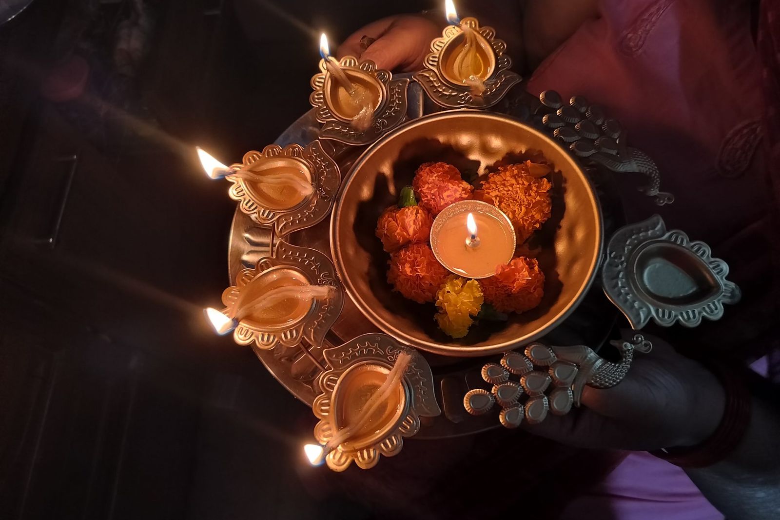 Hands holding a brass aarti plate with lit diyas and marigold flowers